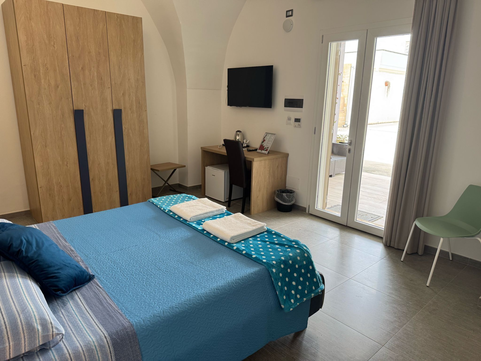 A hotel room with a bed, a desk, a chair, a TV, and a sliding glass door to the outside.