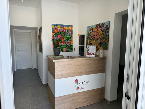 Ein Empfangsschalter mit einem Schild mit der Aufschrift 'Dear Mary', zwei farbenfrohe Blumenbilder und ein Topfpflanze.