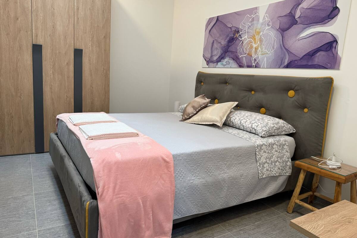 Una cama bien hecha con colcha gris y rosa, dos almohadas y una toalla doblada.
