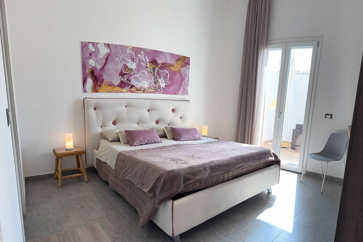 Un dormitorio moderno con una cama grande, una mesita de noche, una silla y un ventanal amplio.