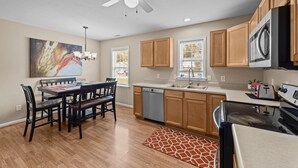 A modern kitchen with wooden cabinets, a dishwasher, a stove, and a dining area with a table and chairs. - 3-Bedroom Retreat with King Suite, Pool Table & Private Fenced Yard (New Bern)