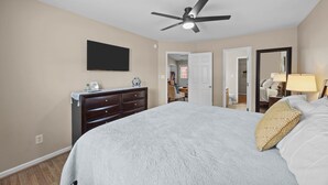 A bedroom with a bed, a dresser, a ceiling fan, a TV, and a door leading to another room. - 3-Bedroom Retreat with King Suite, Pool Table & Private Fenced Yard (New Bern)