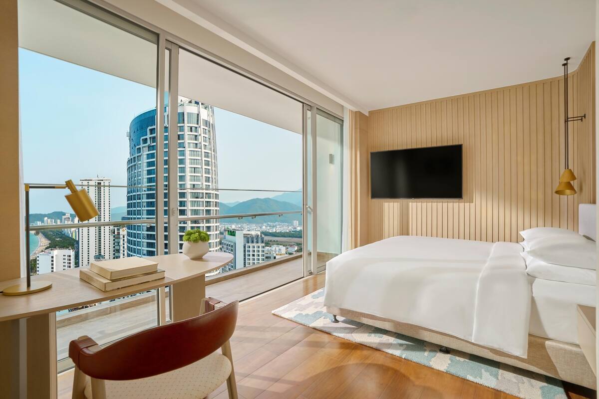 A modern hotel room with a large bed, a desk with a chair, a TV mounted on the wall, and a view of a cityscape and mountains.