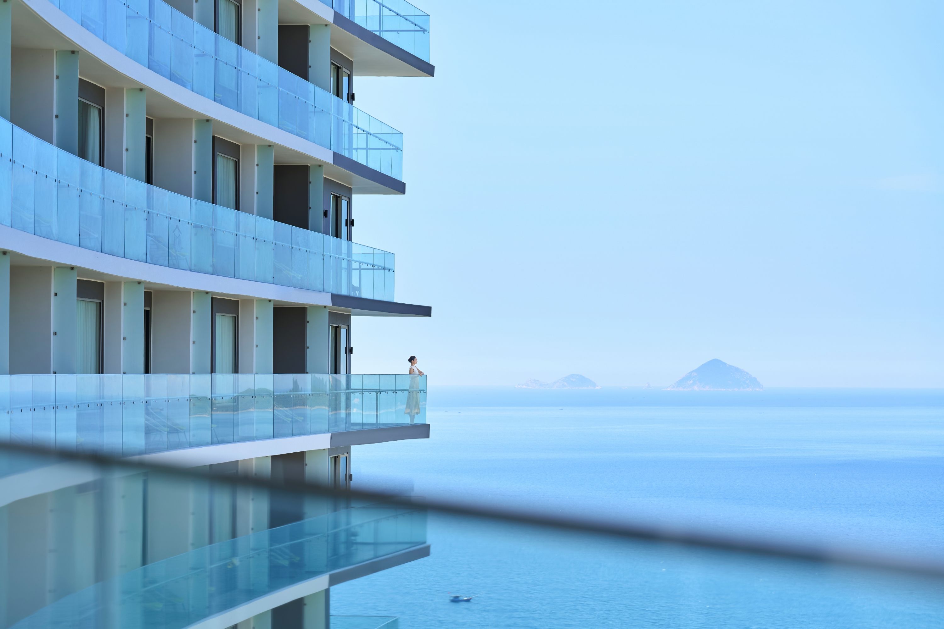 A modern hotel with multiple balconies overlooking a calm sea and distant islands.