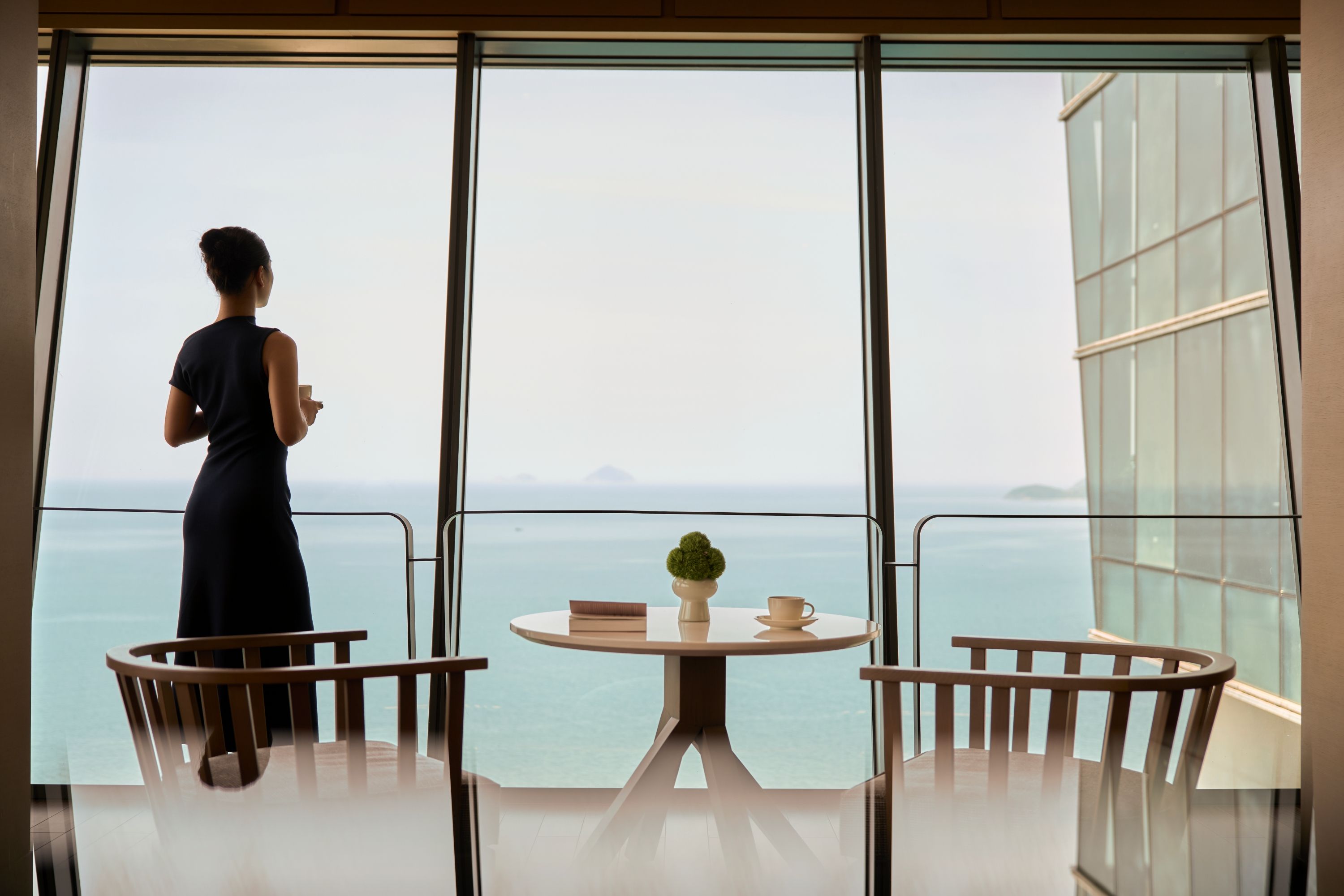 Une femme vêtue d’une robe sombre se tient près d’une fenêtre, d’où l’on aperçoit la mer. Devant elle, une table sur laquelle sont posés une tasse et une petite plante.