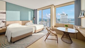 Una habitación de hotel moderna con dos camas, un sofá, una mesa redonda y un amplio ventanal con vistas a la ciudad.