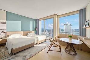 A modern hotel room with two beds, a sofa, a round table, and a large window offering a city view.