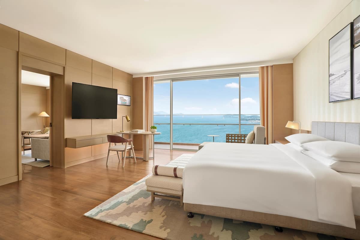A hotel room with a large bed, a desk, a chair, a TV, and a view of the sea.