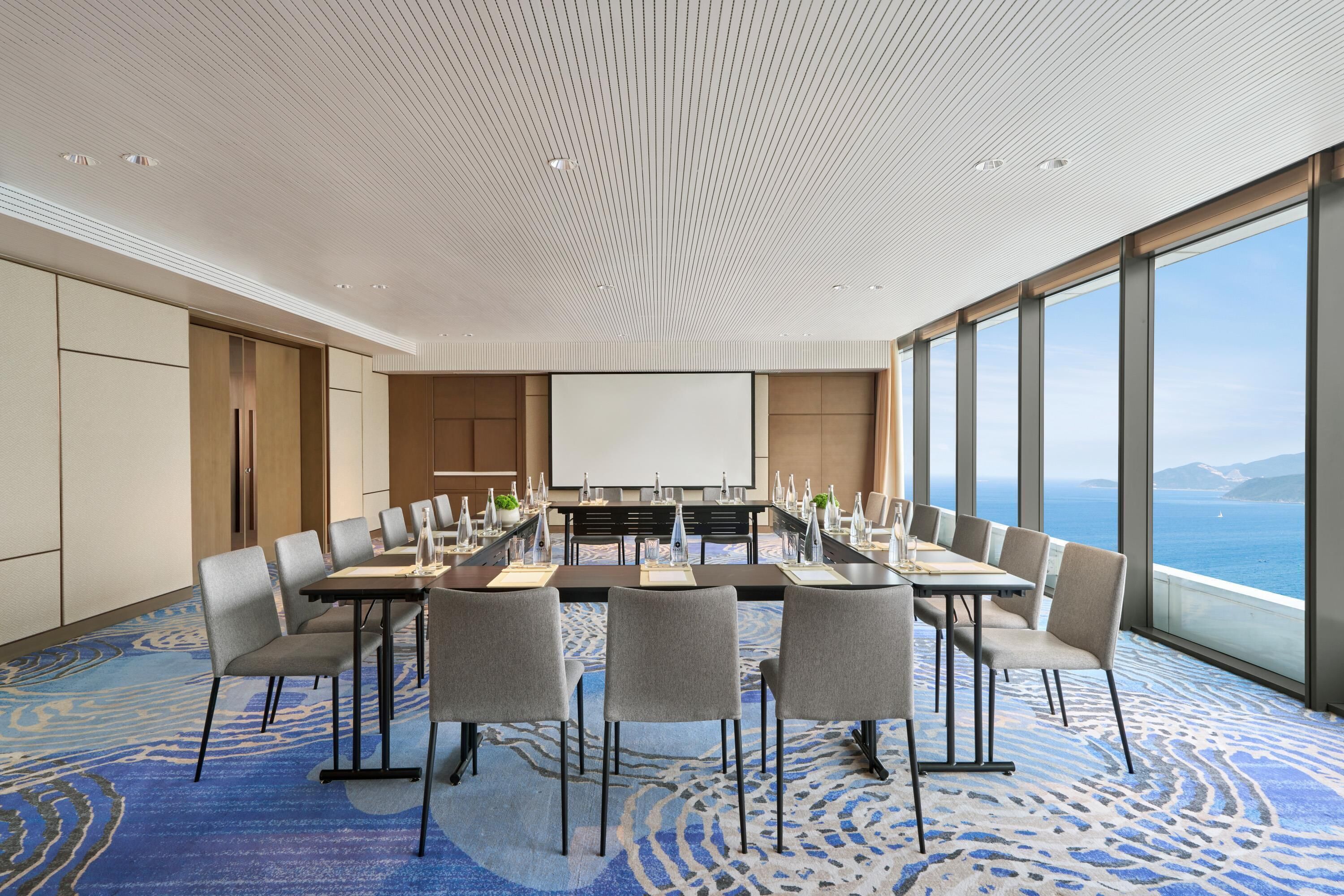 A conference room with a large table, chairs, a projector screen, and a view of the ocean.