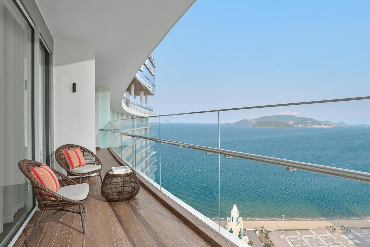 A balcony with wicker chairs, a small table, and a view of the sea and distant land.