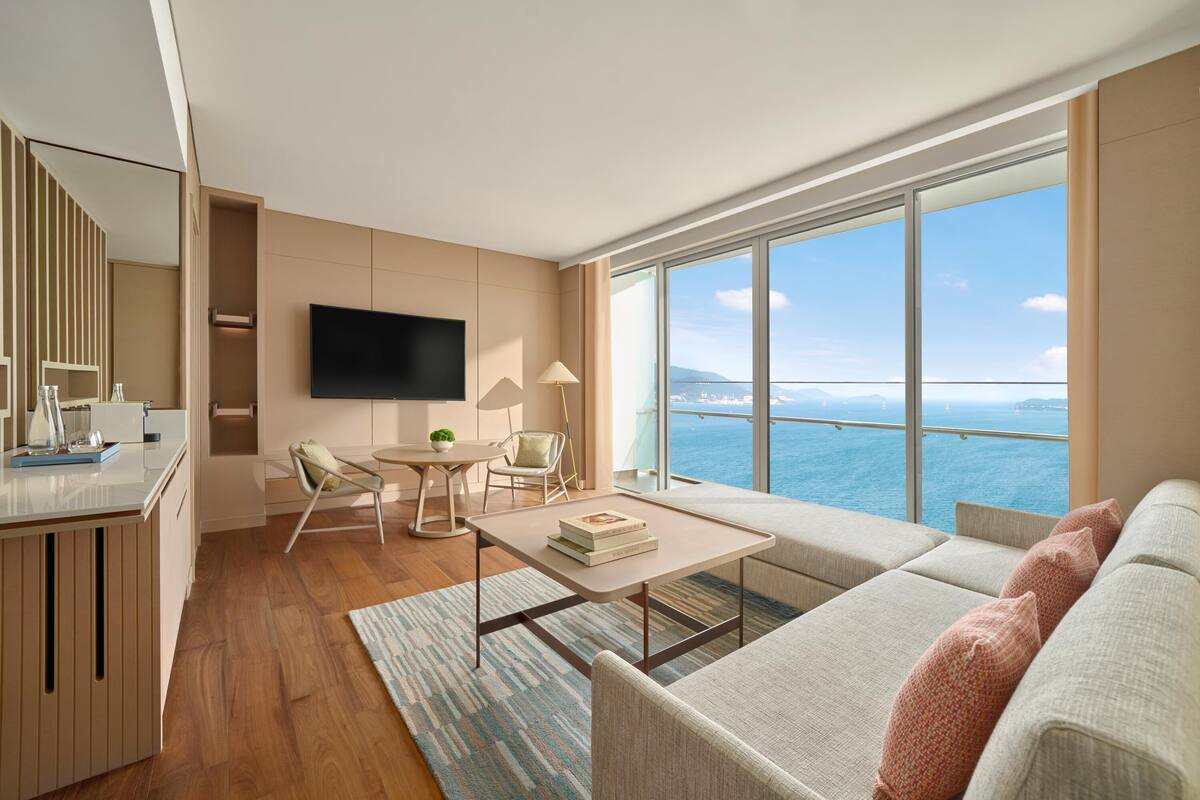 A modern living room with a large window, a flat-screen TV, a dining area, and a sofa with cushions.