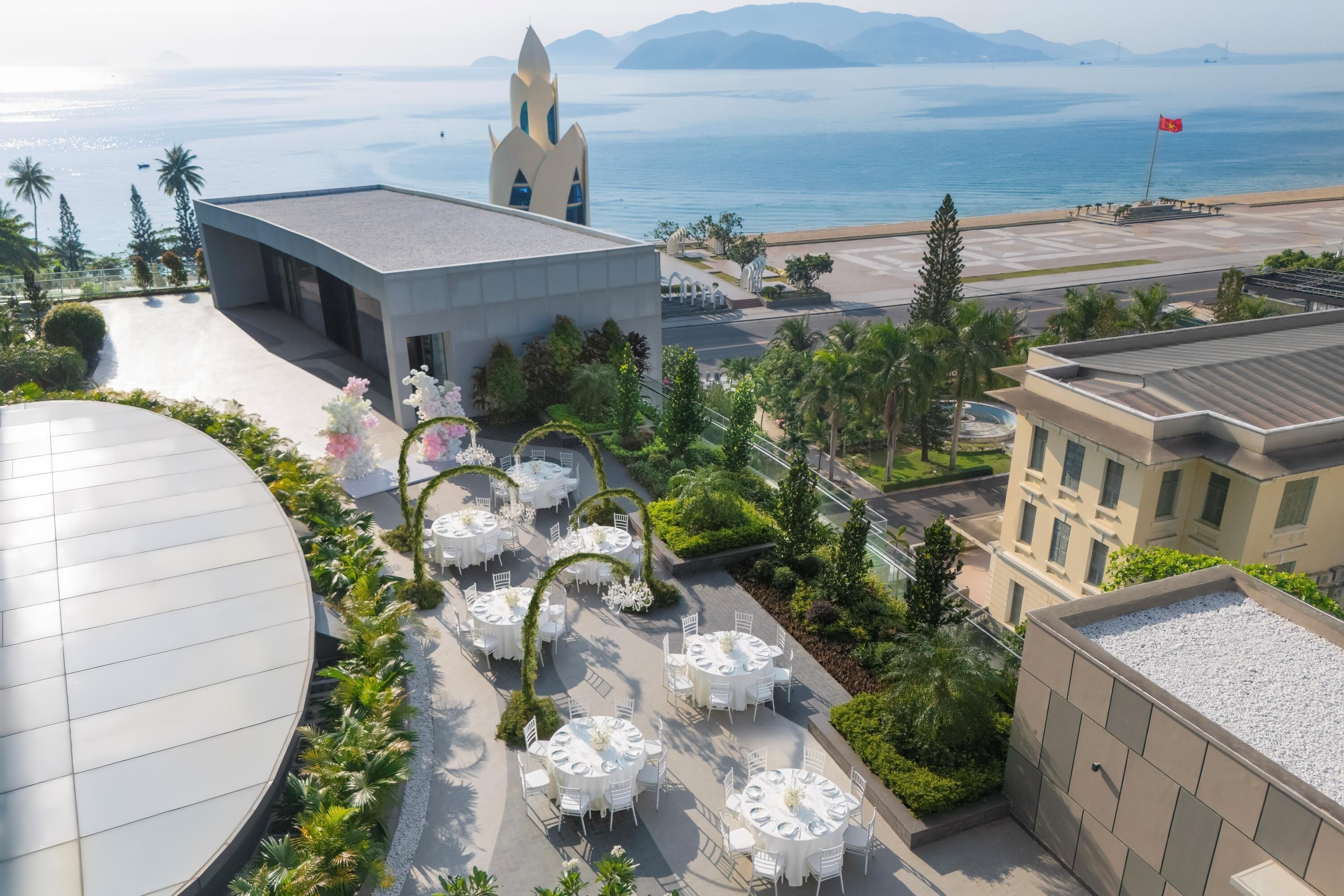 A rooftop terrace with round tables and chairs, surrounded by greenery and overlooking a body of water with a distant mountain range.