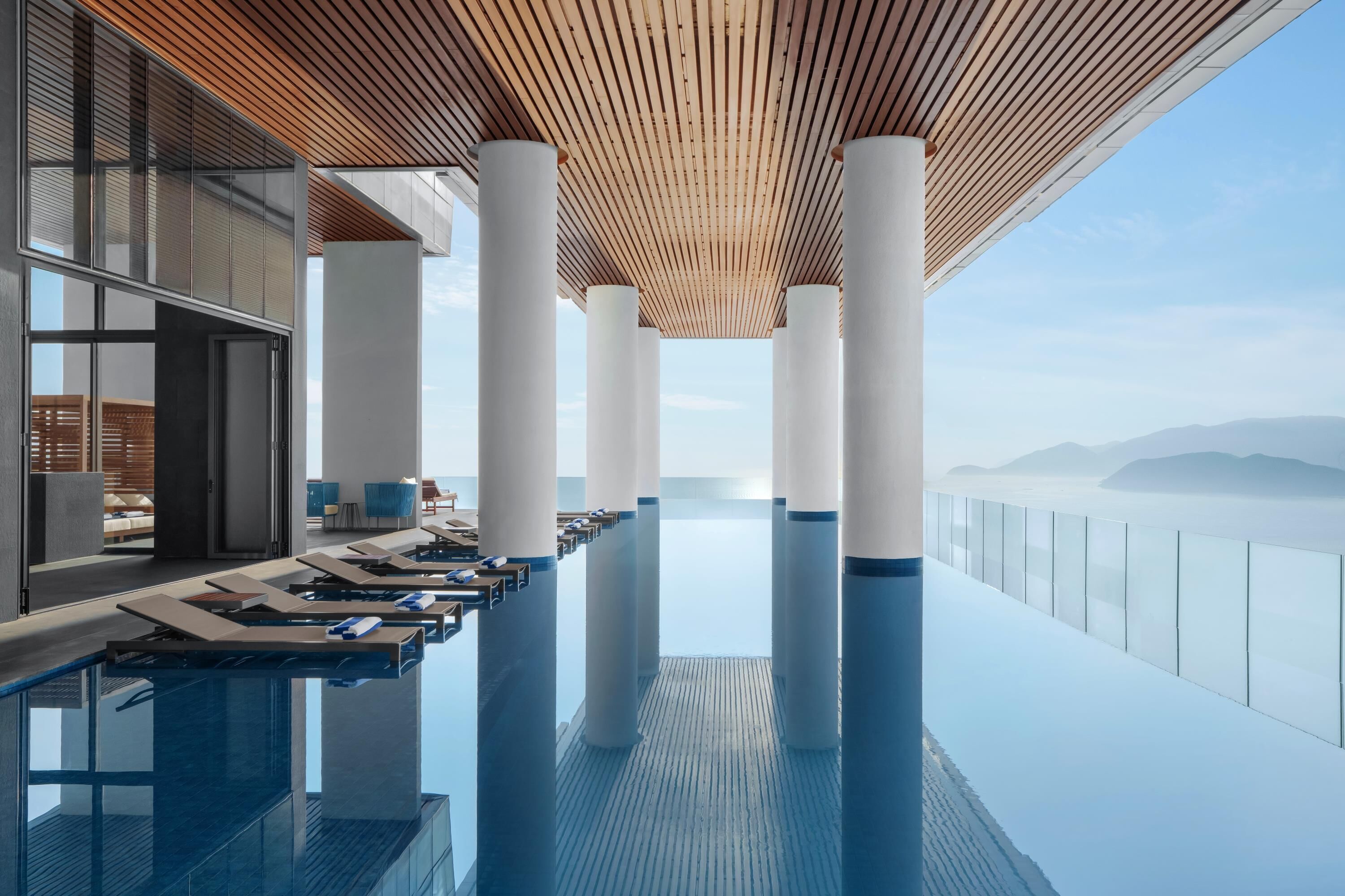 Una piscina infinita moderna con techo de madera y pilares blancos, con vista a un paisaje montañoso.