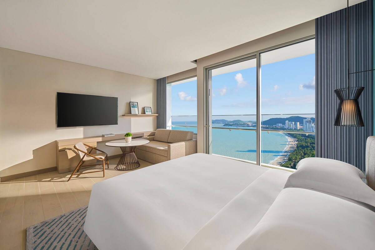 A modern hotel room with a large bed, a seating area with a small table, a flat-screen TV mounted on the wall, and a balcony with a city and beach view.