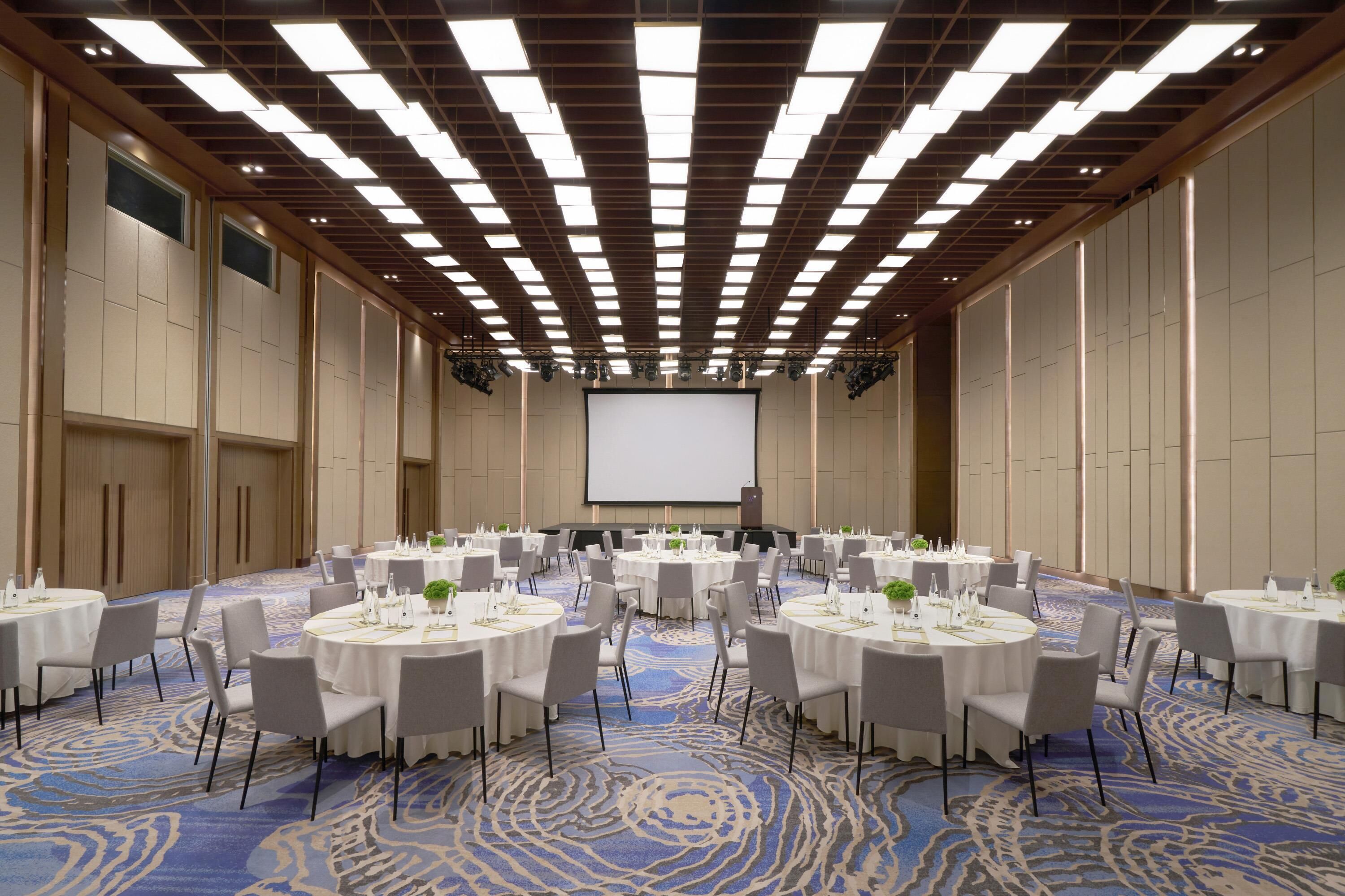 Une salle de conférence équipée de tables rondes, de chaises, d’un écran de projection et d’un tapis à motifs.