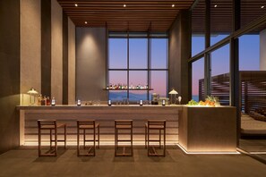 A bar area with a long counter, multiple stools, and a view of the cityscape through large windows. - Hyatt Regency Nha Trang (Nha Trang)