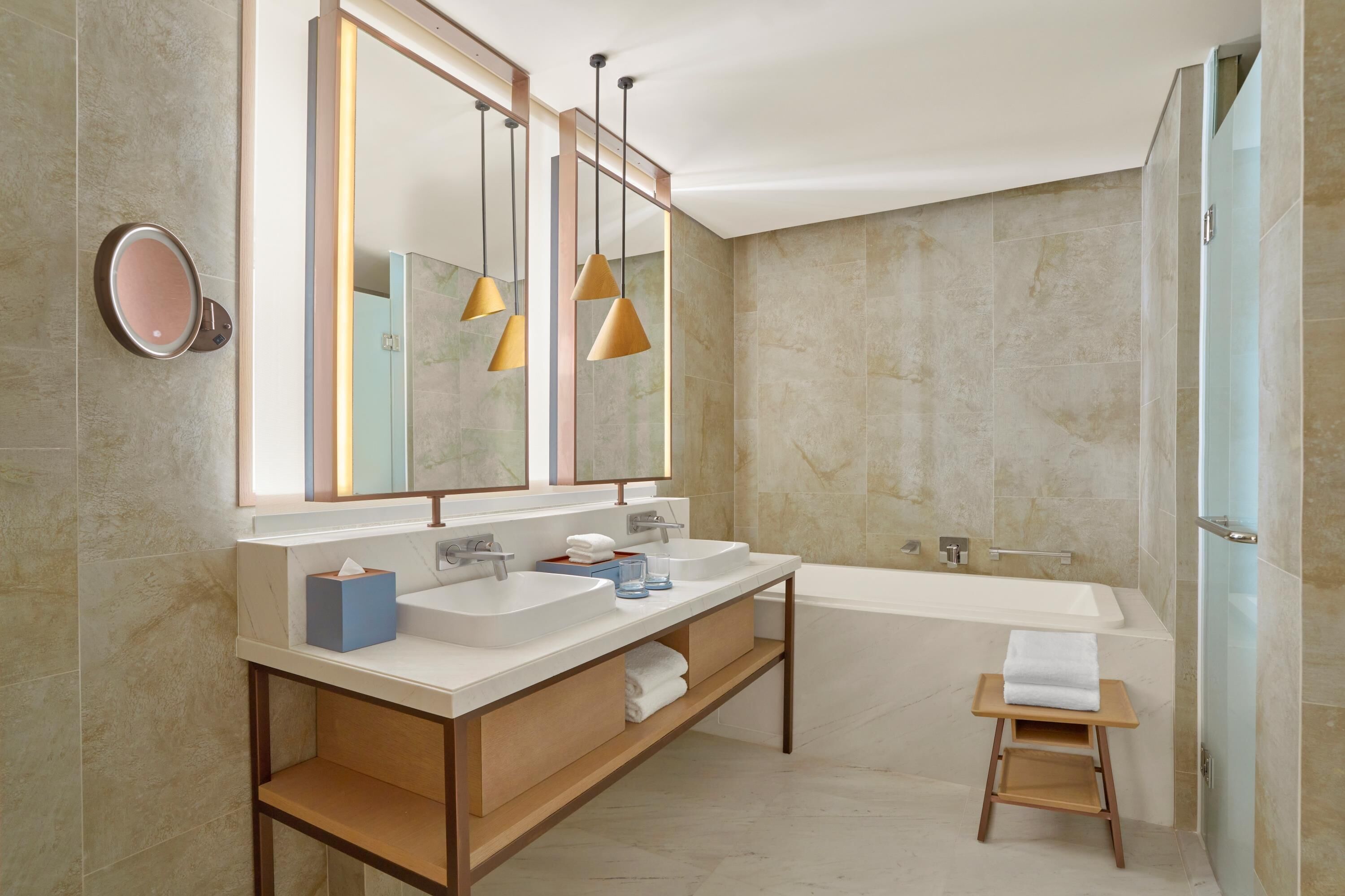 A modern bathroom with a double sink vanity, a large mirror, pendant lights, a bathtub, and a small wooden stool.