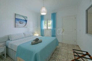 A bedroom with a bed, a painting, a hanging light fixture, and a window with curtains.