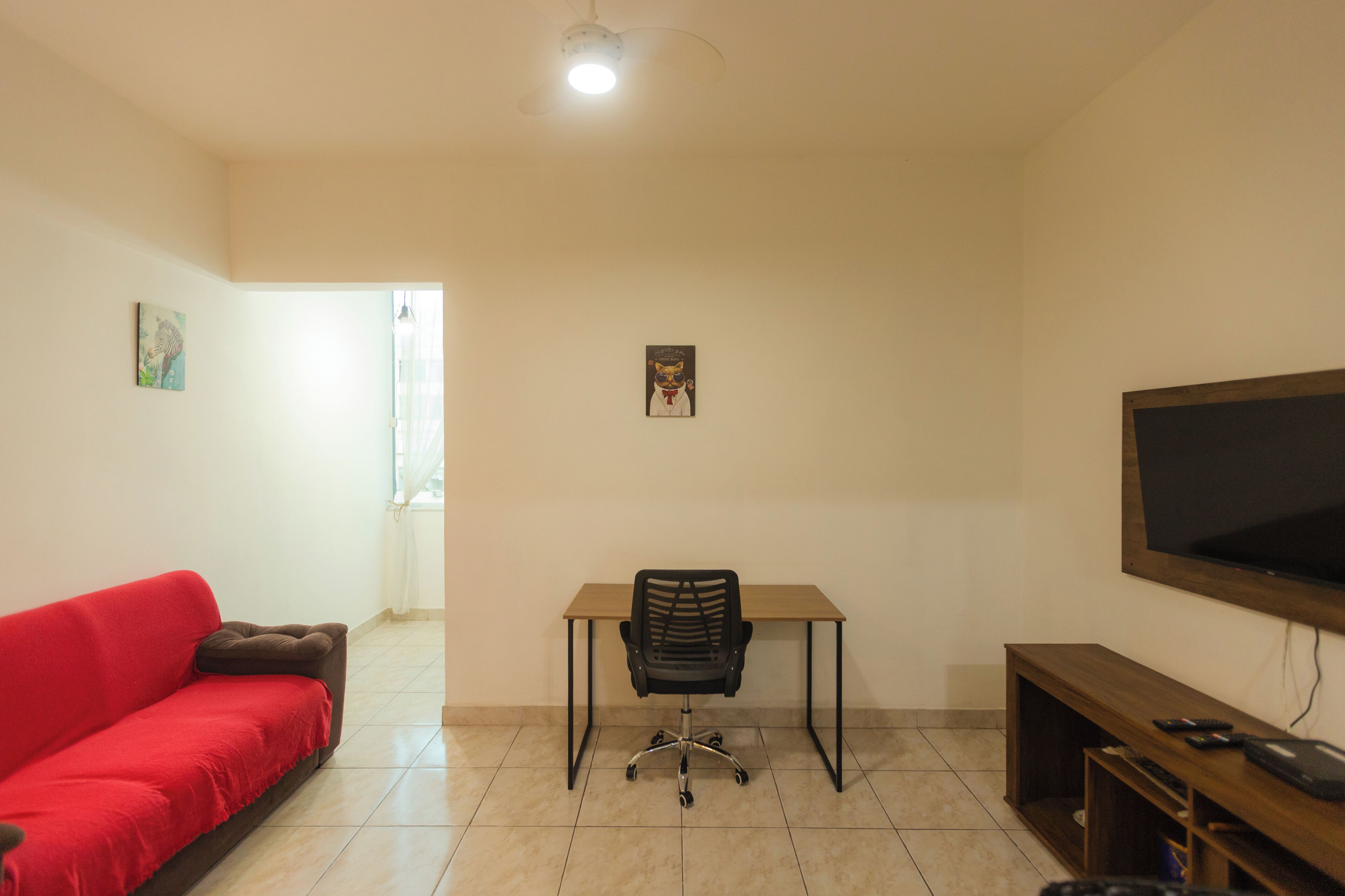 A room with a red sofa, a desk with a chair, a television, and a small wooden shelf.