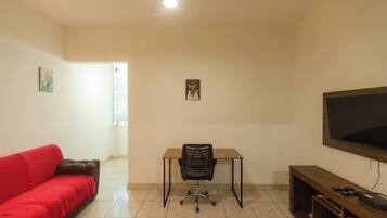 A room with a red sofa, a desk with a chair, a television, and a small wooden shelf.