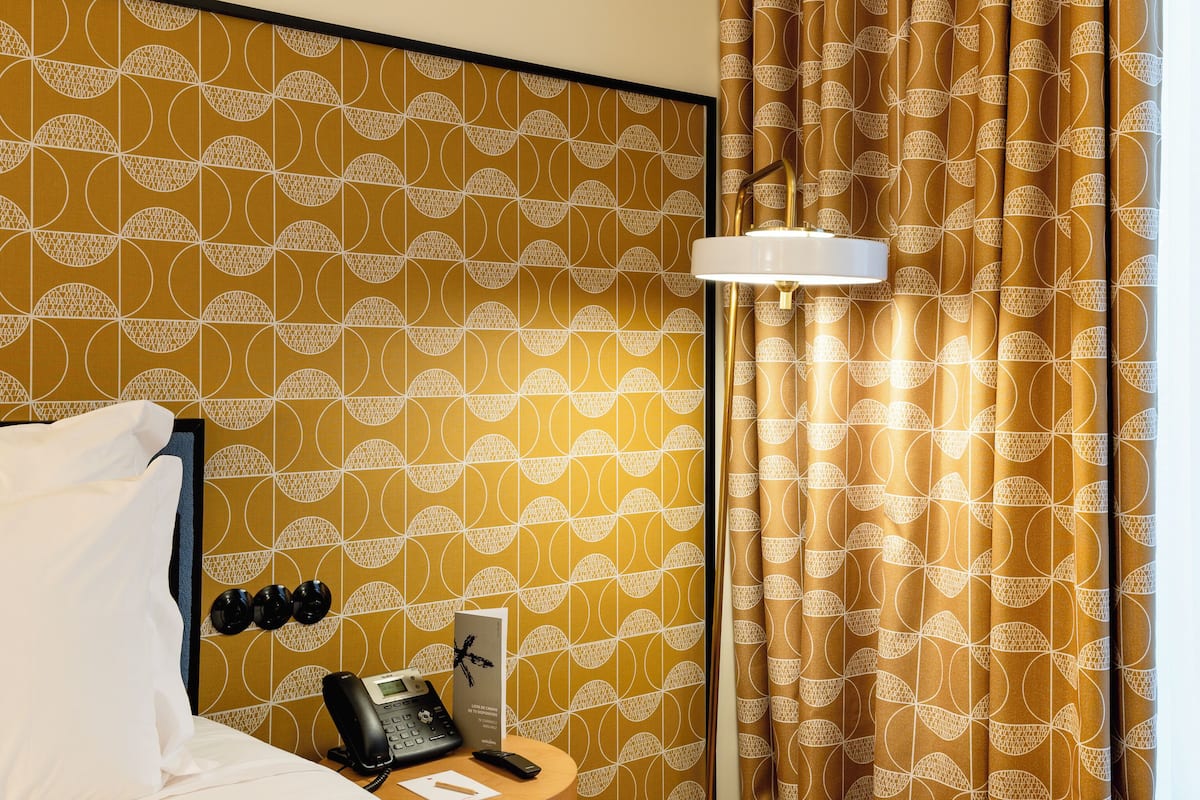 Una habitación de hotel con papel tapiz estampado, una cama con ropa blanca, una mesita de noche con un teléfono y una lámpara, y una cortina con un diseño circular.