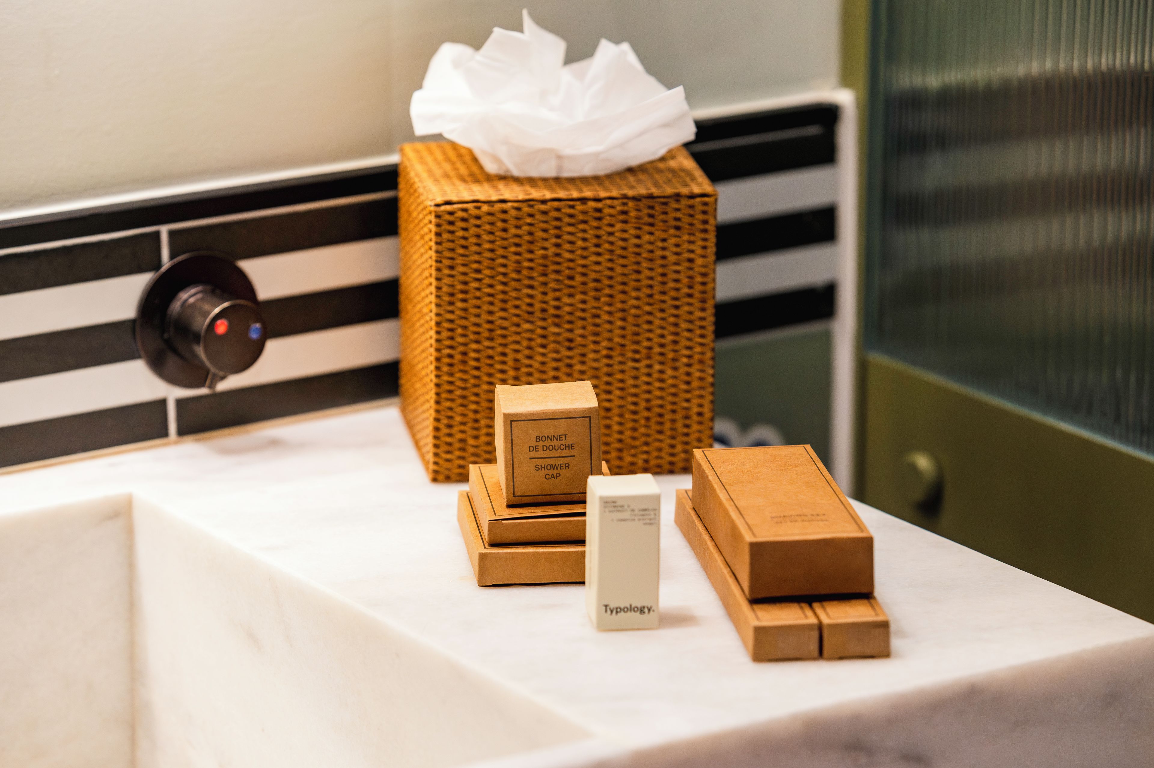 Un baño con un lavamanos blanco, un dispensador de pañuelos de mimbre y tres cajas de madera etiquetadas 'Bonnet de Douche', 'Shower Cap' y 'Typology'.