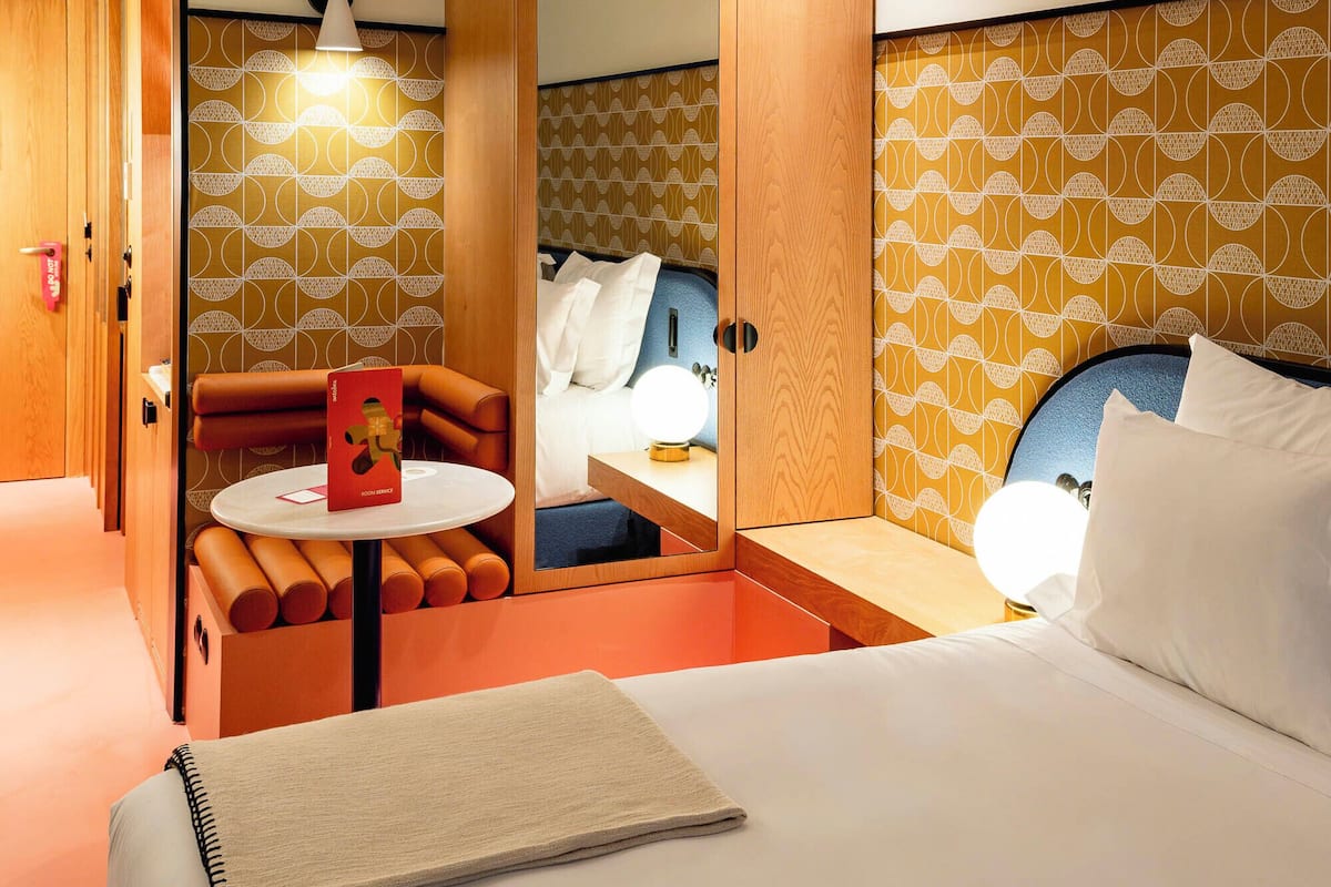 Habitación de hotel con cama, una mesita con un libro, espejo y zona de estar con sofá y sillón.
