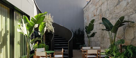 Un área de estar al aire libre con mesas y sillas, rodeada de plantas en macetas y un muro de piedra.