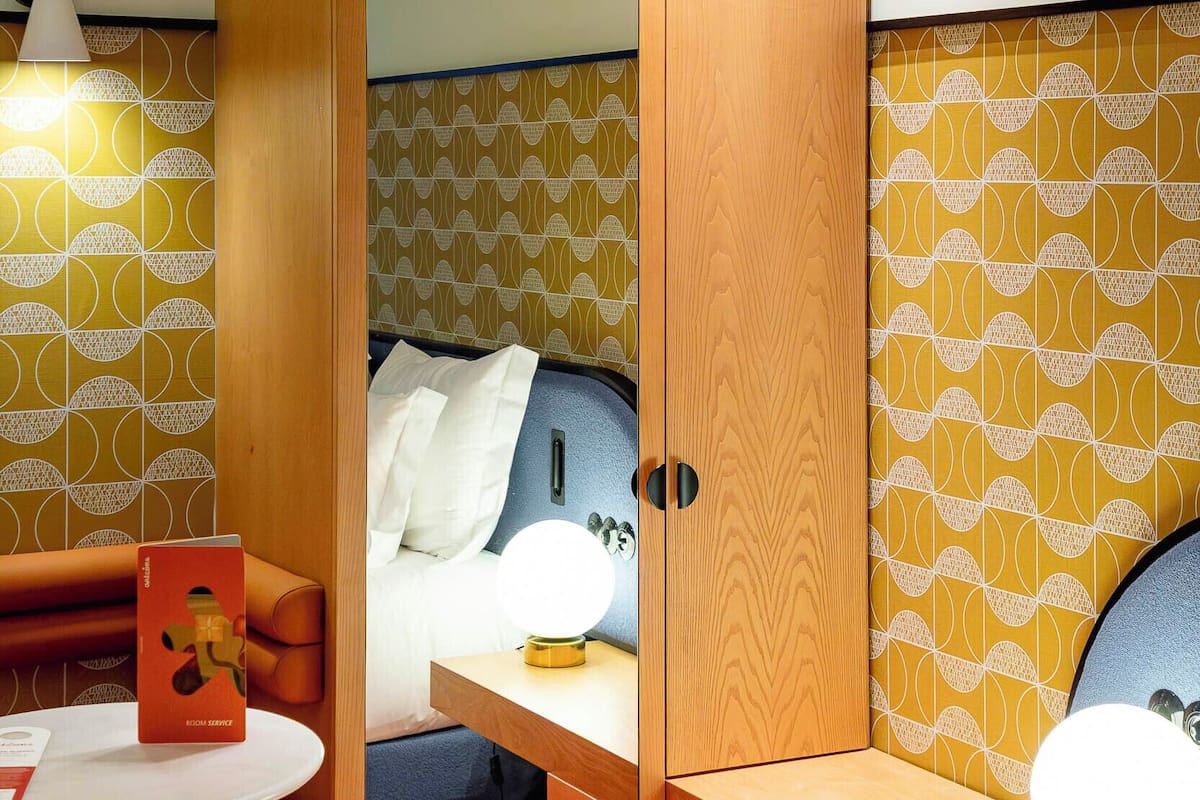 Habitación de hotel pequeña con un armario de madera, una mesita con un libro y una cama con cabecero.