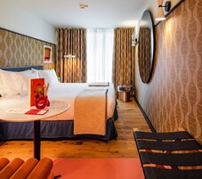 Una habitación de hotel con cama, mesita de noche, silla, espejo redondo y cortinas.