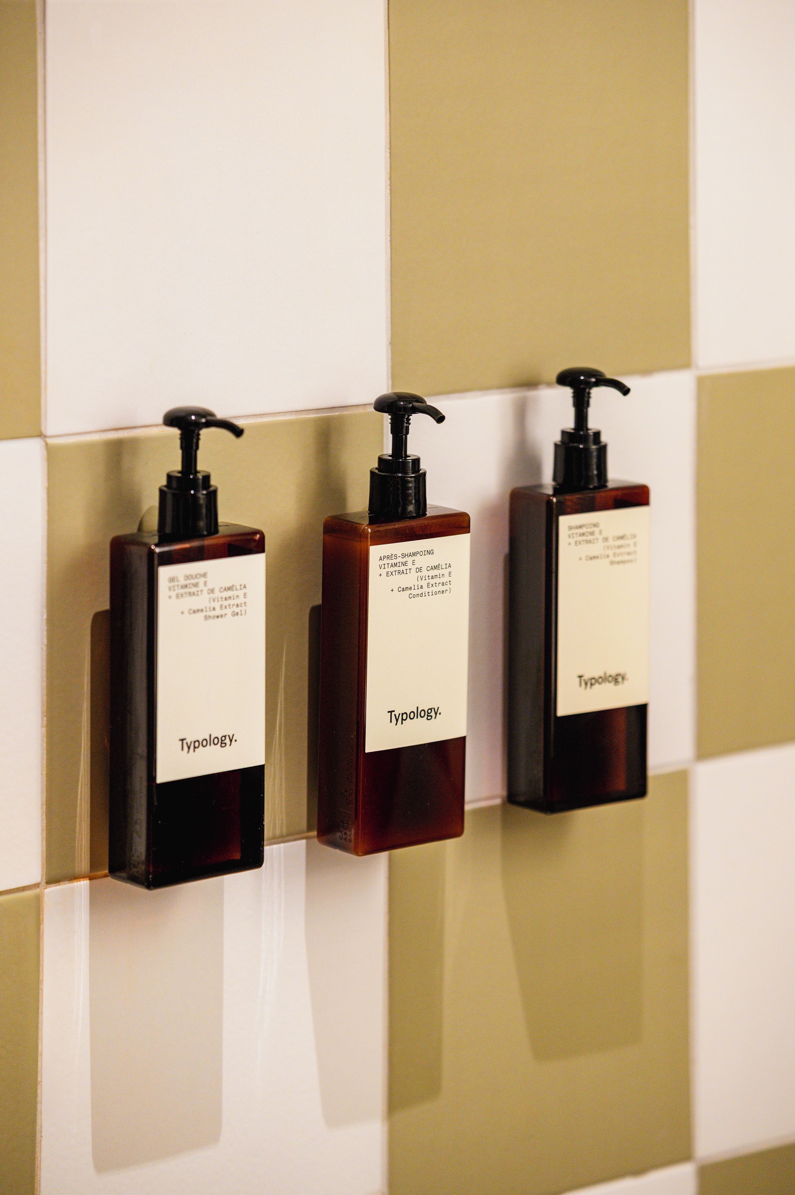 Tres botellas de tocador con dosificadores, cada una con el nombre de una marca, montadas en una pared de azulejos.
