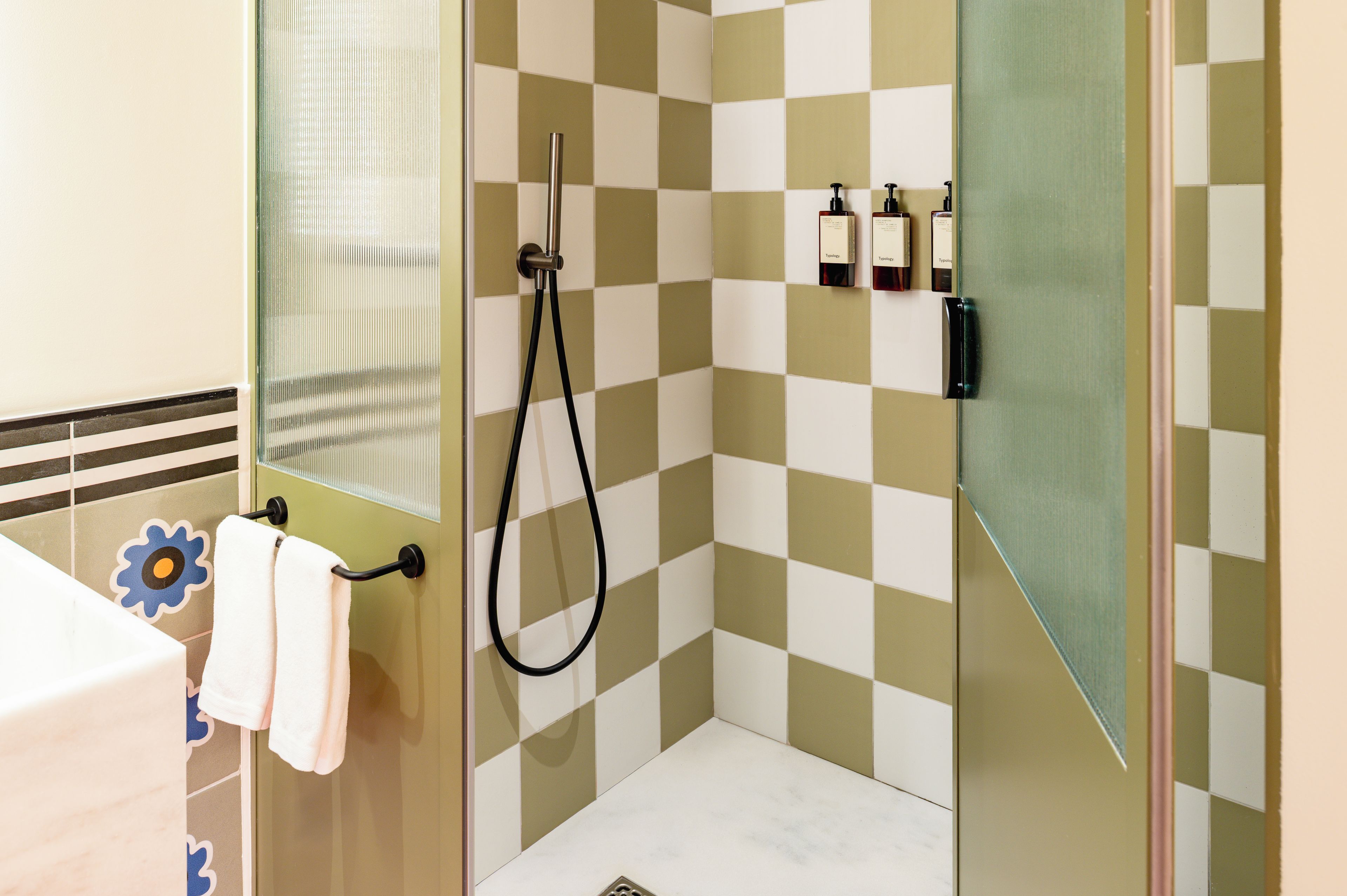 Un baño con pared de baldosas a cuadros, cabina de ducha de vidrio, un perchero con una toalla blanca y un lavabo con dispensador de jabón estampado con flores.