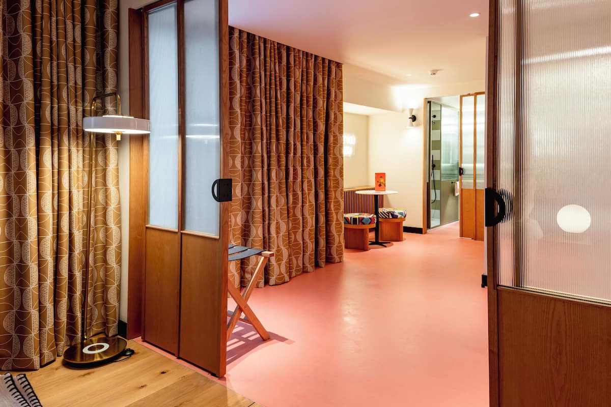 Habitación de hotel moderna con piso de madera, una pared de acento rosa y una puerta corredera hacia el baño.