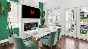 A dining area with a white table set for six, teal chairs, a TV mounted on the wall, and a view through a glass door to the outside. - Elegant Open Concept Close To DC (Arlington)