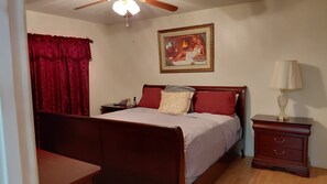 A bedroom with a bed, a nightstand, a lamp, a painting on the wall, and a red curtain. - The Palms at Poinciana: Private Tropical Resort (Kissimmee)