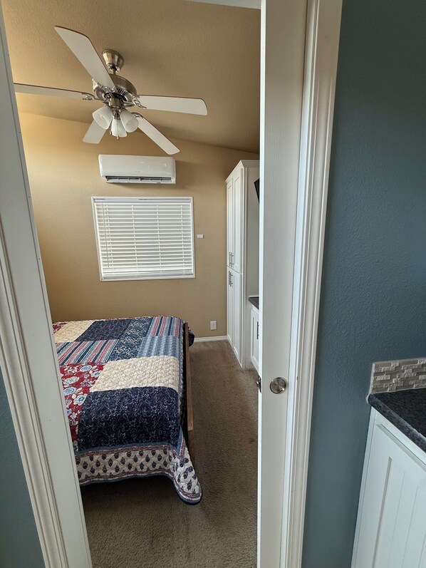 A bedroom with a bed, a ceiling fan, an air conditioning unit, a window with blinds, and a door leading to a bathroom.