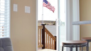 A room with a grey sofa, a wooden deck with a railing, and a view of the beach through a glass door.