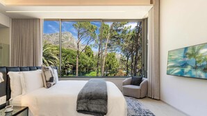 Habitación de hotel moderna con una cama grande, un sofá y un amplio ventanal que ofrece vistas a árboles y montañas.