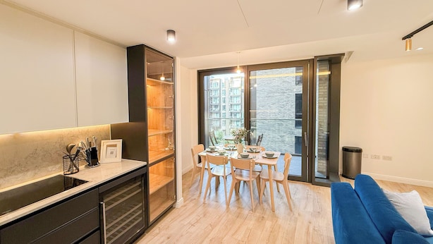 A modern kitchen with a dining area, a blue sofa, and a view of a building through large windows. - Elegant 2-BR Flat w/ Lift & Private Balcony – Farringdon, Clerkenwell, Barbican (London)