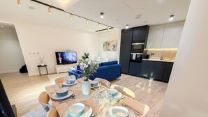 A modern living space with a dining area, a blue sofa, a kitchen, and a TV in the background. - Elegant 2-BR Flat w/ Lift & Private Balcony – Farringdon, Clerkenwell, Barbican (London)