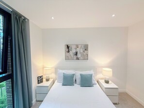 Un dormitorio moderno con una cama grande, dos mesitas de noche con lámparas, una ventana con cortinas y un cuadro abstracto colgado en la pared.