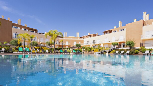 Een zwembad met ligbedden en parasols, omringd door meerlaagsgebouwen met balkons.