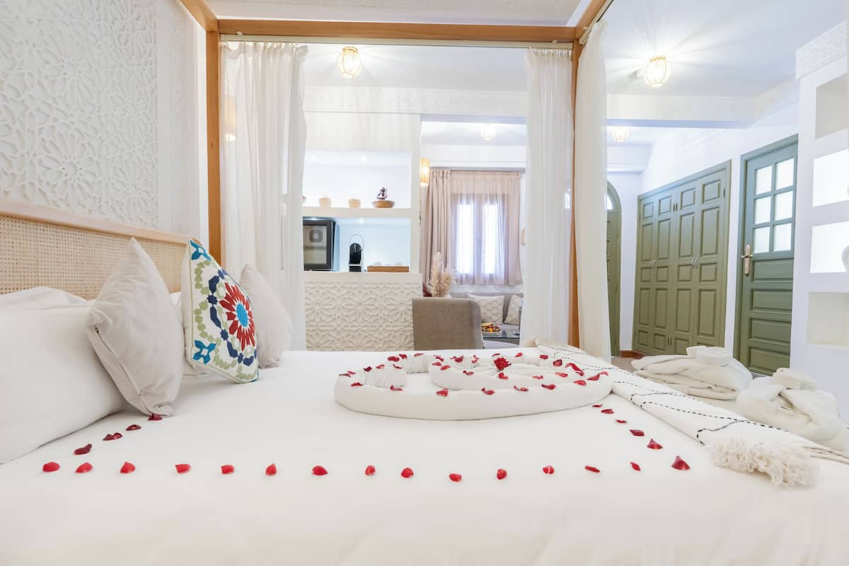 A hotel room with a bed, a small table, a green wardrobe, and a view of the kitchen and dining area.