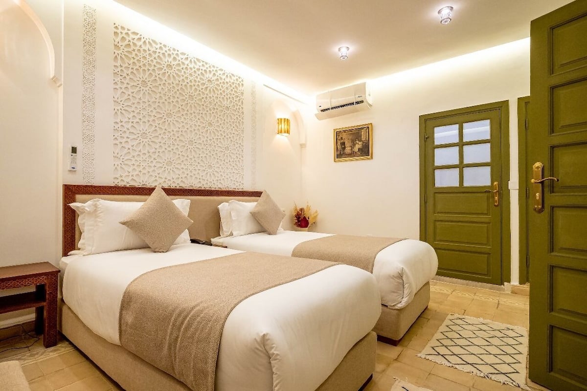 A hotel room with two beds, a decorative wall, an air conditioning unit, a green door, and a small bedside table.
