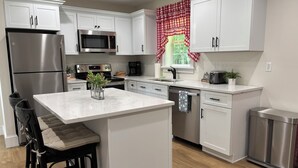A modern kitchen with white cabinets, stainless steel appliances, a central island with bar stools, and a window with red and white checkered curtains. - Cozy Country Cottage in Sullivan County Catskills. 2 Bedrooms, 1 Bath, Heat & AC (Ferndale)