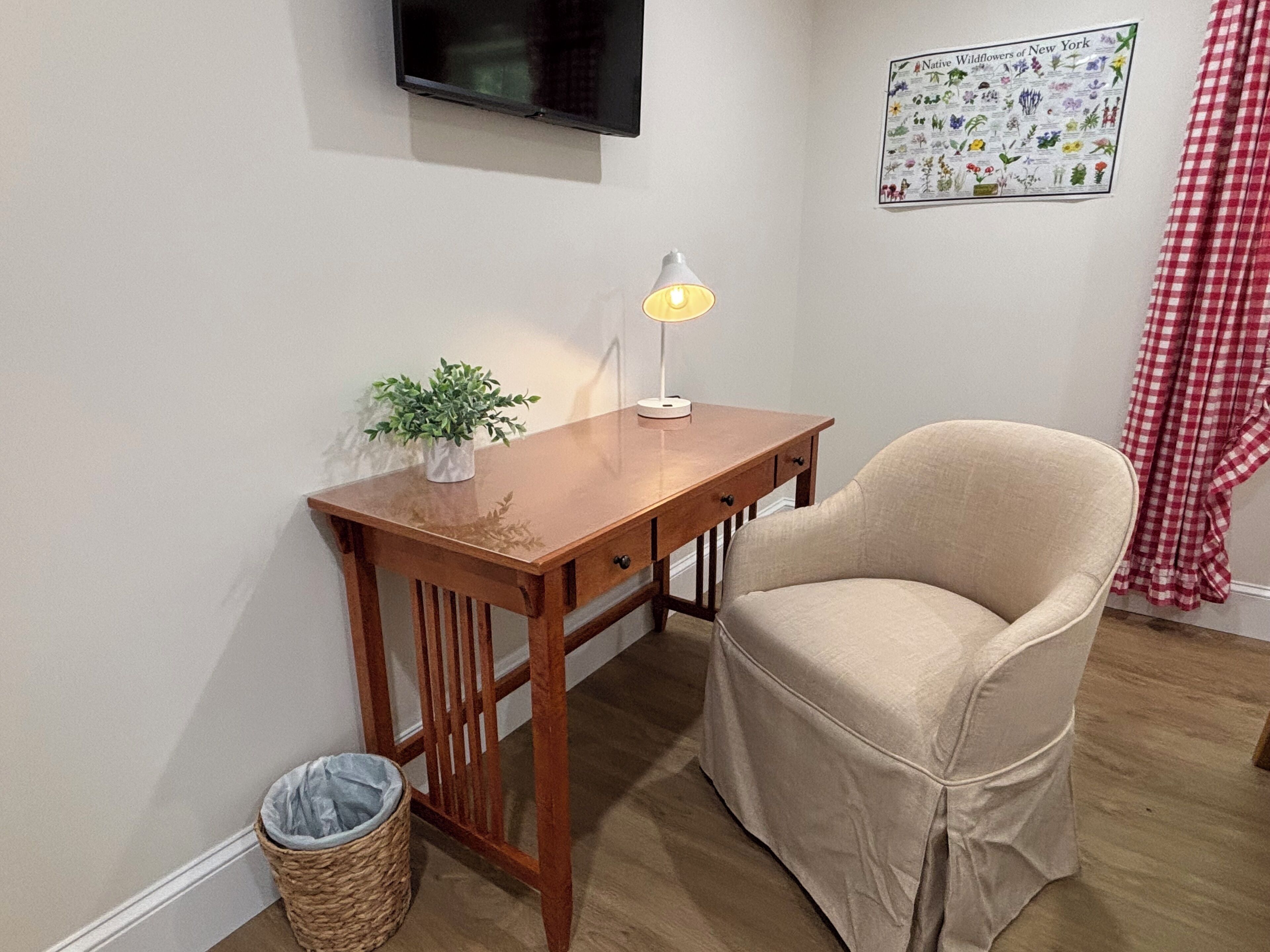 Una habitación pequeña con un escritorio de madera, una silla, una lámpara, un televisor montado en la pared, un cartel y una cesta de mimbre.