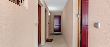 Un pasillo con una puerta roja al final, una lámpara en la pared y una alfombra pequeña en el suelo.
