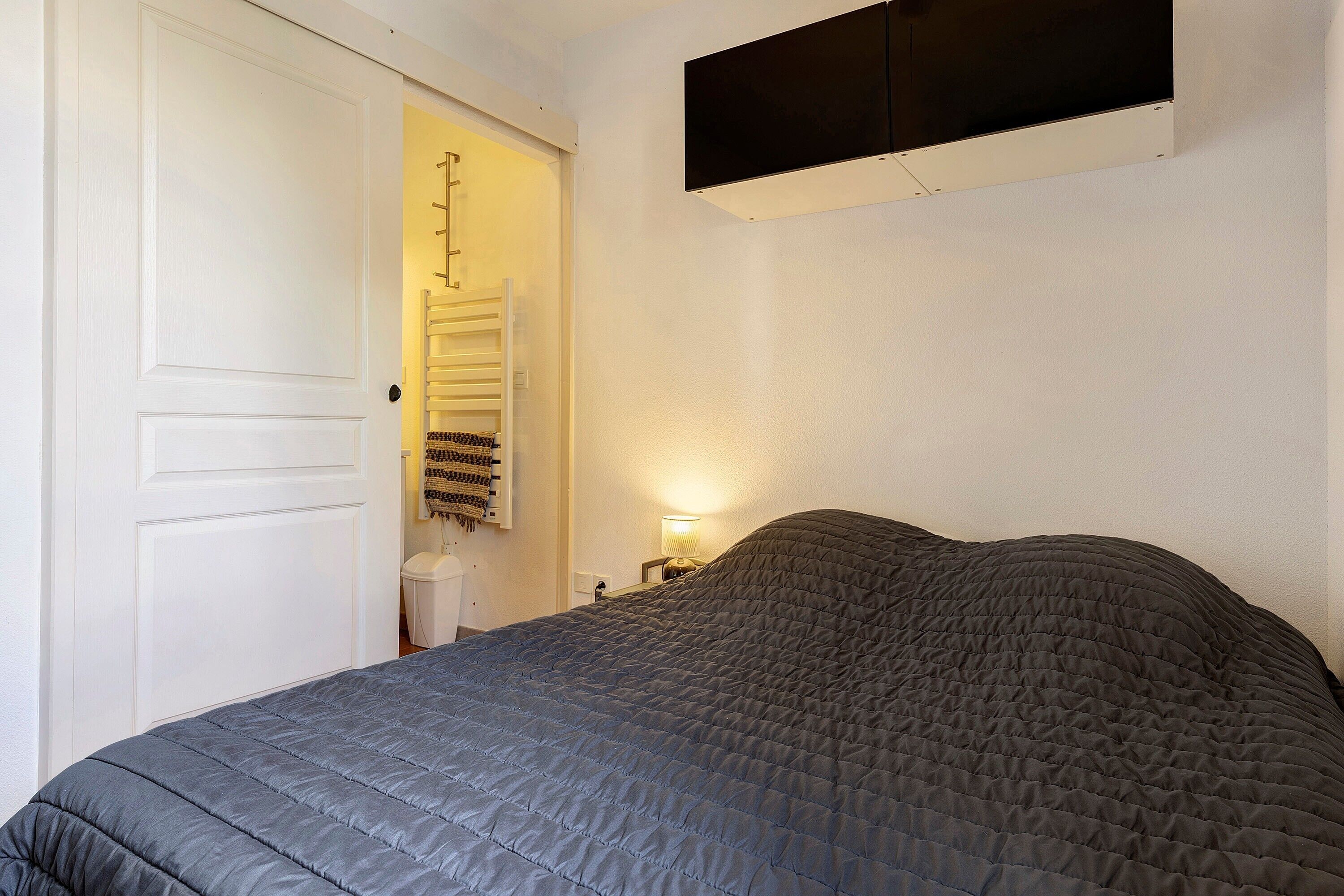 A bedroom with a bed, a door, a wall-mounted shelf, and a bedside table with a lamp.