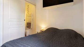 A bedroom with a bed, a door, a wall-mounted shelf, and a bedside table with a lamp.