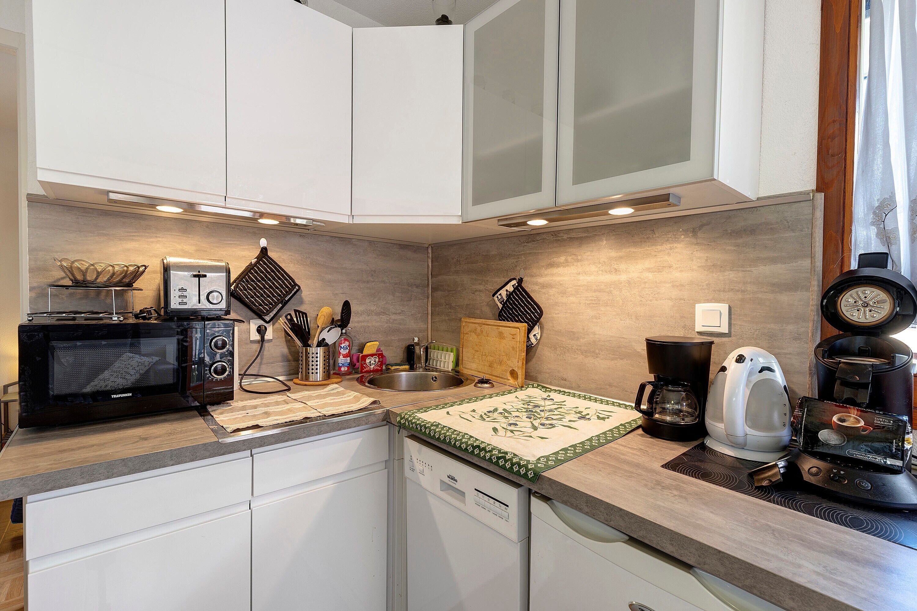 A compact kitchen with white cabinets, a microwave, a toaster, a coffee maker, and a kettle.